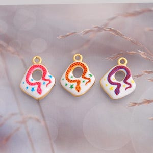 Peut inclure: Trois breloques en métal doré avec des centres en émail blanc, chacune représentant un serpent enroulé et de petites étoiles. Le premier serpent est rose, le deuxième orange et le troisième violet. Chaque breloque a un anneau circulaire.