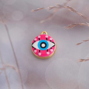 May include: A circular gold-toned metal charm featuring an enamel design of a stylized eye. The eye has a bright blue iris, a black pupil, and a white sclera, set against a vibrant pink background decorated with small black lines and pink dots.