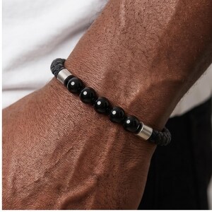 Peut inclure: Bracelet noir à perles avec un bracelet en cuir tressé et des détails en métal argenté. Le bracelet présente une série de perles noires polies. Un accessoire élégant pour un usage quotidien.