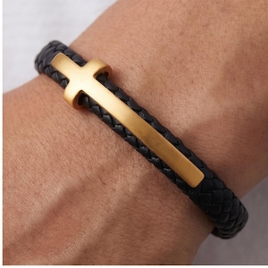 May include: Black braided leather bracelet with a gold-colored cross. The cross is the focal point, set against the dark texture of the woven leather. A simple yet elegant accessory.