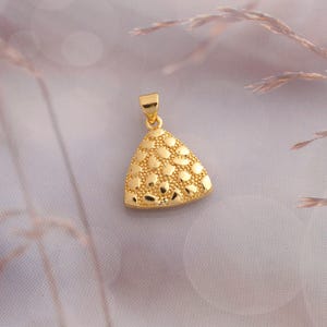 Puede incluir: Un colgante dorado de forma triangular con una superficie texturizada que presenta detalles en relieve redondeados similares a guijarros. El colgante incluye una pequeña anilla triangular lisa en la parte superior.