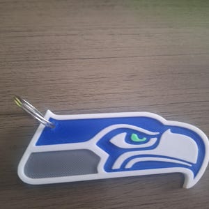 May include: A blue, white, and gray keychain featuring a stylized eagle head design. The keychain has a metal ring for attachment. The eagle head has a green eye and is a team logo.