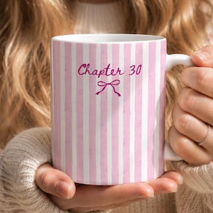 May include: A white ceramic mug featuring vertical light pink stripes and the text Chapter 30 in a dark pink script font, positioned above a small dark pink bow graphic. The mug has a smooth finish and a curved handle on the right side.