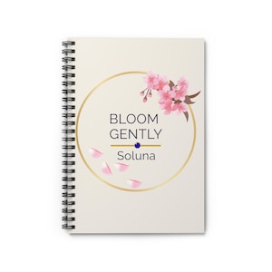 Puede incluir: Cuaderno de espiral de color crema. La portada presenta un círculo dorado con las palabras "BLOOM GENTLY" en azul marino, un punto azul y el nombre "Soluna". Ilustraciones y pétalos de flores de cerezo rosas.