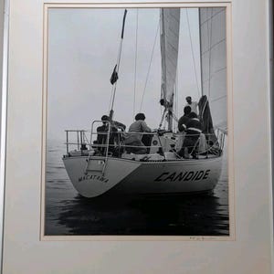 Op de afbeelding: Zwart-witfoto van een zeilboot op het water. De boot, genaamd "Candide" en "Macatawa", heeft verschillende mensen aan boord. De zeilen zijn gehesen en de boot vaart op het water.