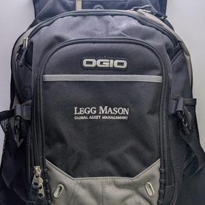 Könnte beinhalten: Ein schwarzer und grauer OGIO-Rucksack mit mehreren Reißverschlussfächern, seitlichen Netztaschen und dem gestickten Text "LEGG MASON GLOBAL ASSET MANAGEMENT" auf der Vorderseite. Das Design umfasst verstellbare Riemen und silberne Akzente.