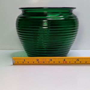 May include: A deep emerald green glass bowl featuring a horizontal ribbed texture and a wide, smooth rim. A yellow measuring tape is positioned in front, indicating the bowl has a diameter of approximately 5.5 inches at its widest point.