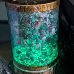 May include: A cylindrical diffuser with a faux wood grain finish on the top and bottom. The central transparent chamber is filled with small, polished green and brown stones, illuminated from within by a bright green light. Two buttons labeled LIGHT and MIST are visible on the base.