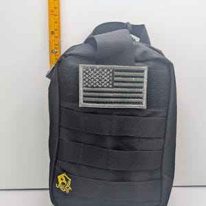 May include: A black tactical pouch with a grey American flag patch and MOLLE webbing. A yellow USCCA logo is on the bottom left. A yellow measuring tape is positioned vertically along the left side, showing the pouch is approximately 8 inches tall.