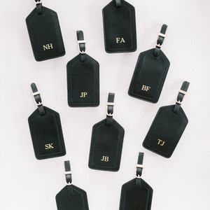 May include: A collection of nine black rectangular luggage tags with gold-toned buckles and straps. Each tag features unique gold-embossed initials including NH, FA, JP, BF, SK, JB, TJ, JD, and DB, arranged in a scattered pattern on a light background.