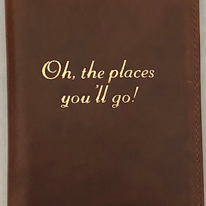 Pu&ograve; includere: Un portapassaporto in pelle marrone con scritta dorata "Oh, the places you'll go!"