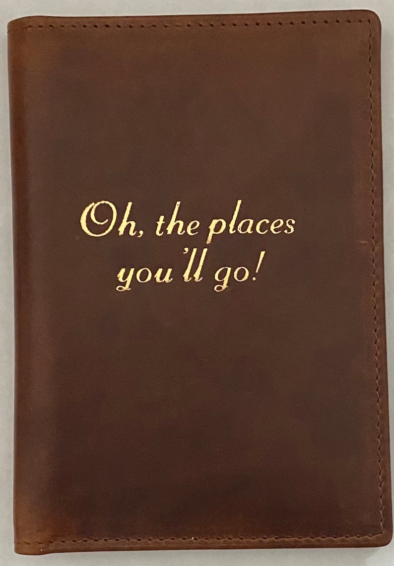 Pu&ograve; includere: Un portapassaporto in pelle marrone con scritta dorata "Oh, the places you'll go!"