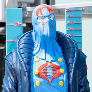 May include: A blue and red bust of a character with a mask and a jacket. The bust is approximately 18 cm tall. The character has a red cobra emblem on the chest and yellow buttons.
