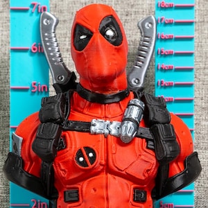 May include: A red and black Deadpool bust figure featuring two silver swords on the back, black tactical gear, and a circular chest emblem. The figure is positioned against a blue measuring tool showing a height of approximately 18 cm.