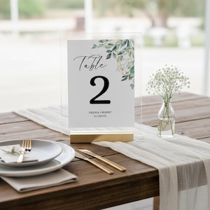 Puede incluir: Una tarjeta con el número de mesa de boda con el número 2 en negrita negra. La tarjeta tiene un diseño floral y los nombres Valeria y Robert con una fecha. La tarjeta está en un soporte acrílico transparente con base dorada.