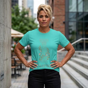 Op de afbeelding: Tealkleurig T-shirt met een gouden illustratie van een klassieke zuil en bloemendetails. Het woord "AMPHIS" staat eronder gedrukt. Het shirt met korte mouwen is gemaakt van zacht materiaal.