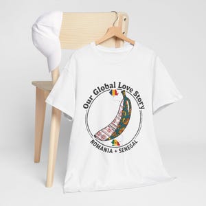Könnte beinhalten: Weißes T-Shirt mit einem Bananen-Design mit Mustern und dem Text "Our Global Love Story". Das Bananen-Design integriert die Flaggen von Rumänien und Senegal. Eine weiße Baseballkappe liegt auf einem Holzstuhl.
