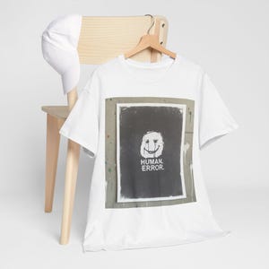 Puede incluir: Camiseta blanca con un estampado gráfico de una cara sonriente distorsionada y las palabras "HUMAN. ERROR." Una gorra de béisbol blanca descansa sobre una silla de madera. La camiseta está colgada en una percha de madera.