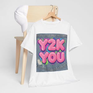 Könnte beinhalten: Weißes T-Shirt mit dem Text "Y2K YOU" in rosa Buchstabenblasen, vor einem Jeans-Taschen-Design. Das Shirt hängt an einem Holzkleiderbügel. Eine weiße Baseballkappe liegt auf einem Holzstuhl.
