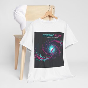 May include: White t-shirt with a graphic design featuring the text "COSMIC FLUX" and "REBIRTH IN COLOR." The design showcases a swirling vortex of teal and magenta colors against a dark background. A white baseball cap rests on a wooden chair.
