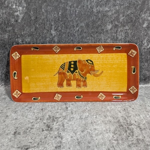 May include: A rectangular ceramic tray featuring a central yellow panel with a brown elephant illustration wearing a decorative blanket. The border is a deep reddish-brown colour, accented with repeating diamond-shaped floral motifs and small green paisley shapes.