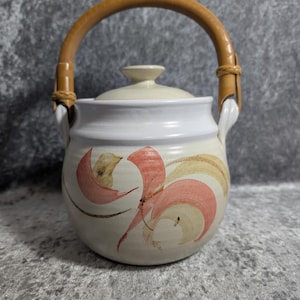 May include: A white ceramic teapot with a lid and a woven handle. The teapot features a floral design in shades of pink, brown, and yellow. The handle is made of woven light brown material. The teapot is a decorative kitchen item.
