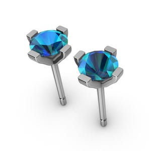 Könnte beinhalten: Ein Paar Ohrstecker mit runden, facettierten blauen Edelsteinen, die in einem silberfarbenen Metall gefasst sind. Die Edelsteine haben ein mehrfarbiges Aussehen mit Blau- und Türkistönen. Die Ohrringe werden vor einem weißen Hintergrund präsentiert.