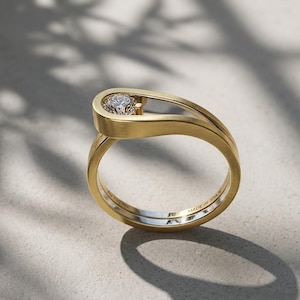 May include: A yellow gold ring with a brushed matte finish featuring a single round brilliant-cut diamond set within an open, curved top section. The inner band is engraved with the text AU 750 and MADE IN SPAIN, showcasing a sleek design.