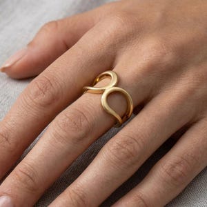 May include: A gold-toned ring featuring an open, fluid design with two interconnected, rounded loops that create a continuous, smooth shape. The metal has a soft, matte finish, and the band is structured with a curved, organic silhouette.