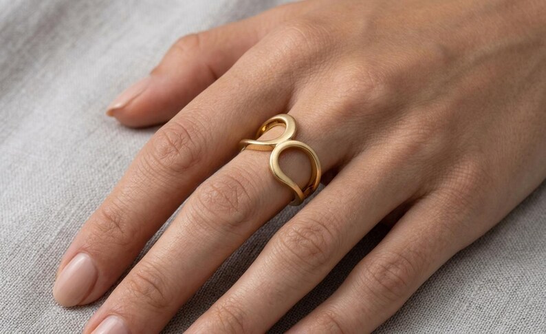 May include: A gold-toned ring featuring an open, fluid design with two interconnected, rounded loops that create a continuous, smooth shape. The metal has a soft, matte finish, and the band is structured with a curved, organic silhouette.