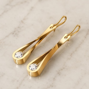 Può includere: Un paio di orecchini pendenti color oro con un design in metallo ritorto a forma di goccia. Ogni orecchino è impreziosito da una singola pietra preziosa trasparente a taglio rotondo alla base, fissata con una semplice chiusura a gancio.