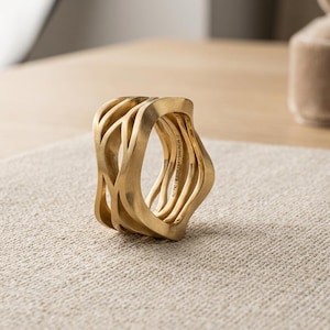 May include: A wide gold ring featuring a multi-layered, openwork design with fluid, wave-like bands. The surface has a matte, brushed finish. The interior of the band is engraved with the text AU 750, 5324, and ARTISAN PIZA 12.