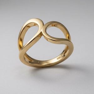 May include: A polished gold ring featuring a continuous, fluid band that loops to create two open, rounded shapes. The metal surface is smooth and reflective, with a hallmark stamp visible on the inner side of the band, showcasing a clean design.