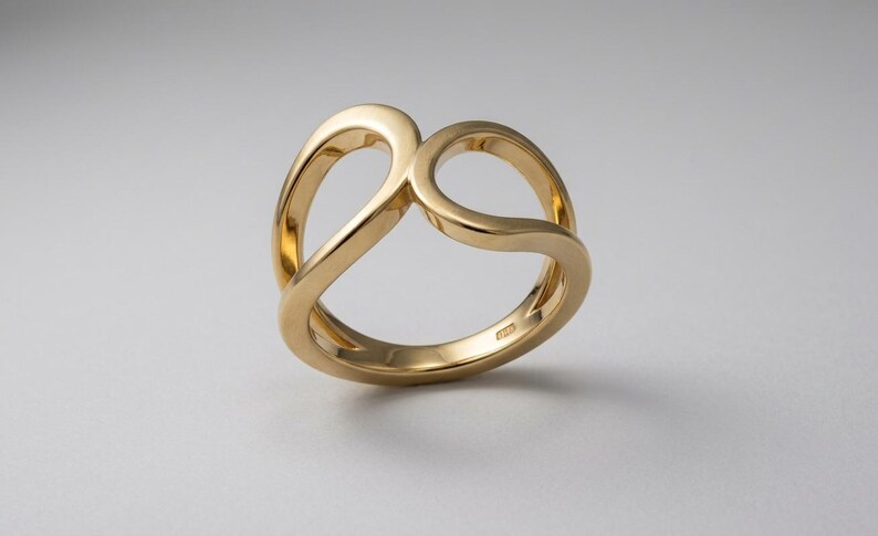 May include: A polished gold ring featuring a continuous, fluid band that loops to create two open, rounded shapes. The metal surface is smooth and reflective, with a hallmark stamp visible on the inner side of the band, showcasing a clean design.
