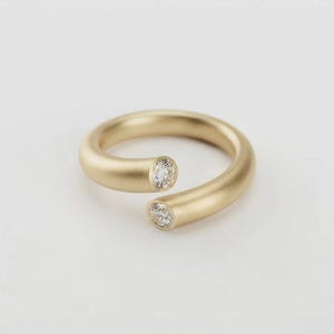 May include: A bypass style ring crafted from matte yellow gold, featuring a smooth, rounded band. The two open ends of the ring are each set with a single, round, clear, faceted gemstone, positioned to face each other with a small gap between them.