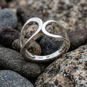May include: A polished sterling silver ring featuring an open, intertwined loop design. The band is stamped with the 925 hallmark on the interior surface, indicating the silver content. The ring has a smooth, reflective finish throughout its structure.