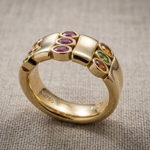 May include: A gold ring with the hallmark AU750 stamped on the inner band. The ring features a series of oval-cut gemstones in shades of red, orange, and green, set between smooth, polished gold segments along the top of the band.
