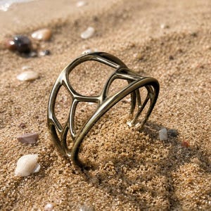May include: A metallic gold-toned cuff bracelet featuring an openwork design with interconnected, branch-like structures. The piece has a smooth, polished finish and a curved, circular shape that is partially embedded in a sandy surface.