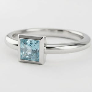May include: A silver-toned ring featuring a square-cut, light blue gemstone set in a smooth bezel setting. The band is polished and rounded, providing a clean, reflective surface that complements the faceted, pale blue stone at the center.
