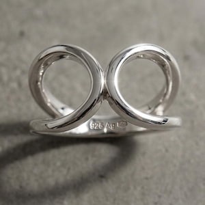 May include: A polished sterling silver ring featuring two interconnected circular loops that rise from a solid band. The inner surface of the band is clearly stamped with the hallmark 925 Ag, indicating the silver content of this metallic jewelry piece.