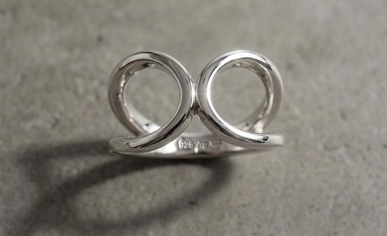 May include: A polished sterling silver ring featuring two interconnected circular loops that rise from a solid band. The inner surface of the band is clearly stamped with the hallmark 925 Ag, indicating the silver content of this metallic jewelry piece.