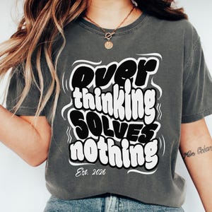 Puede incluir: Camiseta gris oscuro con la frase "Overthinking Solves Nothing" en un diseño audaz en blanco y negro. La camiseta también incluye "Est. 2026". Una prenda casual.