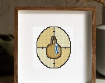 May include: A framed cross-stitch artwork featuring a target design with a central object. The artwork uses shades of tan, brown, and blue. The frame is made of wood.