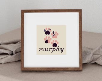 May include: A framed cross-stitch featuring paw prints in shades of pink and purple, with the name "murphy" stitched below. The artwork is set against a cream background and framed in a dark wood.