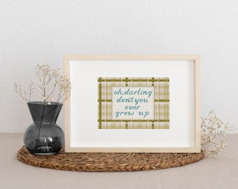May include: A framed print with the text "oh, darling don't you ever grow up" in teal script. The print has a plaid pattern in shades of green and tan. A dark gray vase with dried flowers sits to the left of the frame.