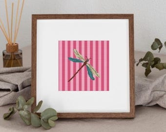 May include: A framed cross-stitch artwork featuring a dragonfly. The dragonfly has blue, green, and brown details, set against a pink and white striped background. The frame is a warm brown color, adding a classic touch to the piece.