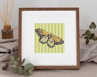 May include: A framed cross-stitch art piece featuring a monarch butterfly. The butterfly has orange, black, and white details, set against a green and white striped background. The frame is a warm brown color.