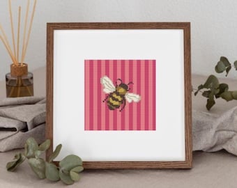 May include: A framed cross-stitch art piece featuring a bee design. The bee is yellow and black with white wings, set against a pink and red striped background. The frame is a warm brown color, and the artwork is displayed on a neutral surface.