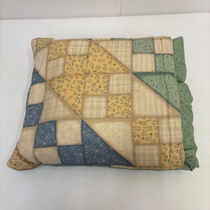 May include: A patchwork cushion featuring a mix of yellow, blue, and green fabrics. The cushion has a quilted design with floral and plaid patterns. The edges are a solid green fabric with a ruffled trim.