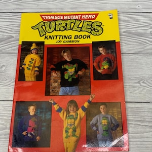 May include: A vintage knitting book titled "Teenage Mutant Hero Turtles Knitting Book" with images of children wearing knitted turtle-themed sweaters. The book has a yellow and red cover with the title in large, green letters.